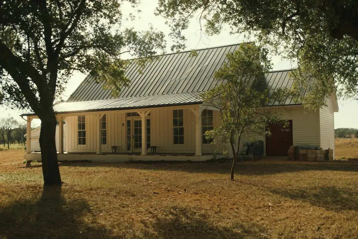 A white board-and-batten barndominium home with a standing-seam metal roof and a covered porch under live oaks