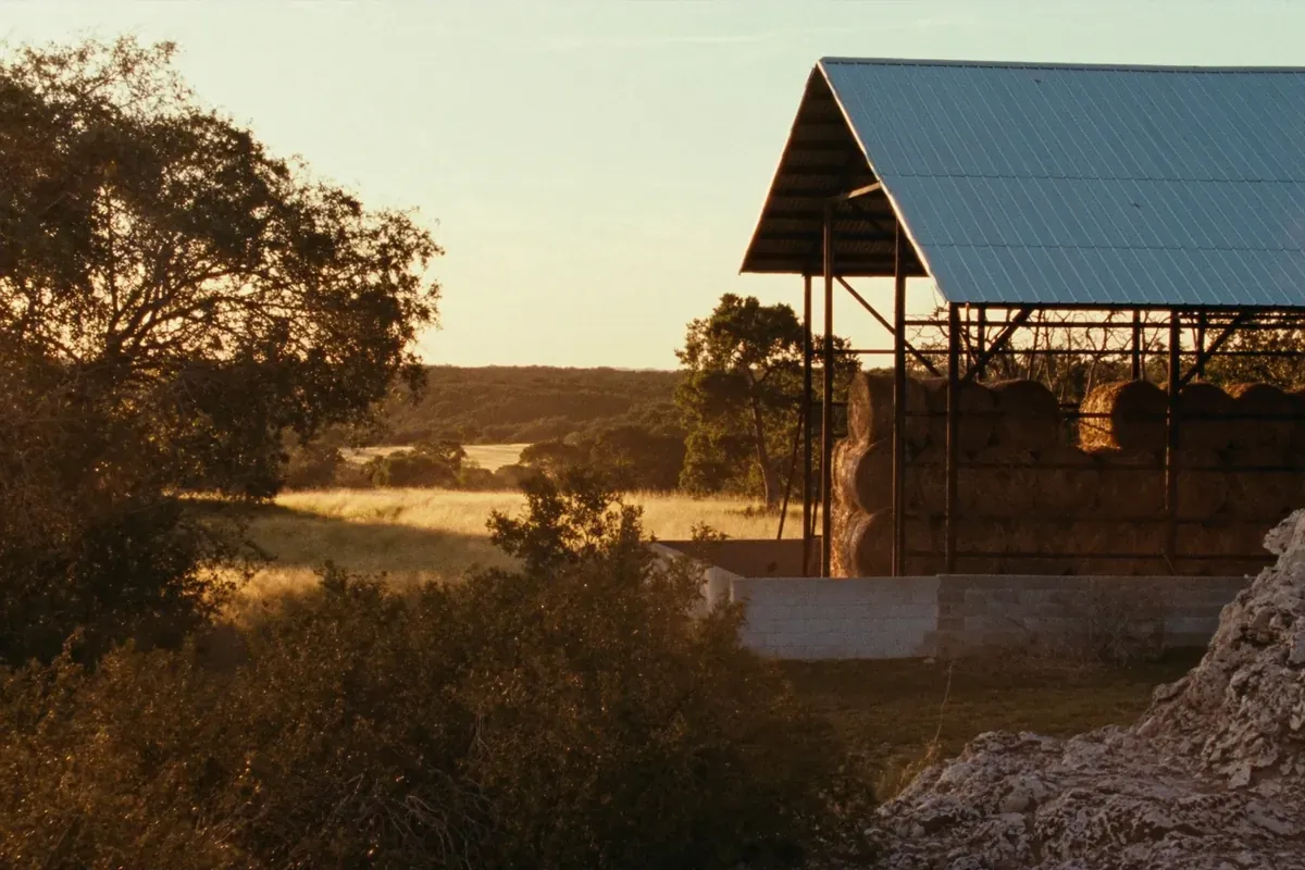 A steel-frame hay barn stacked with round bales on Hill Country ranch land at golden hour