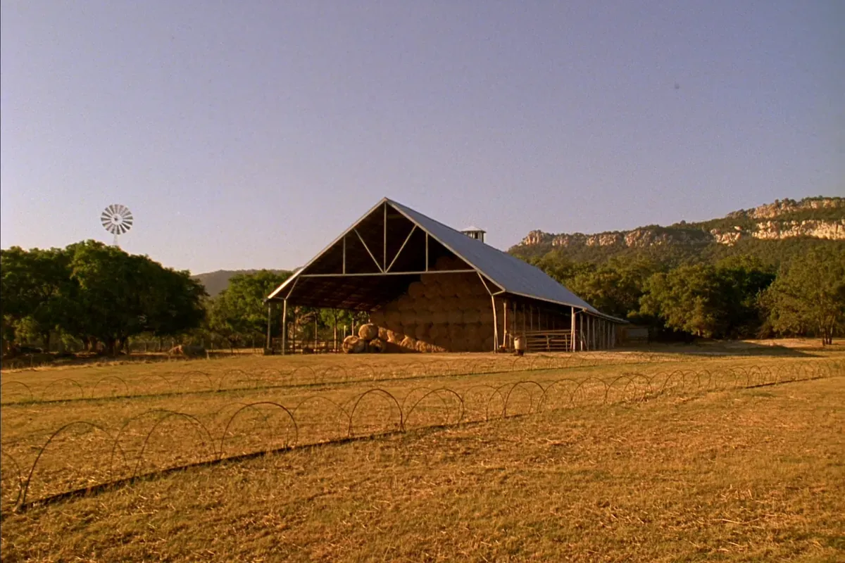 An open hay barn stacked with round hay bales beside a windmill and a limestone bluff in the Texas Hill Country