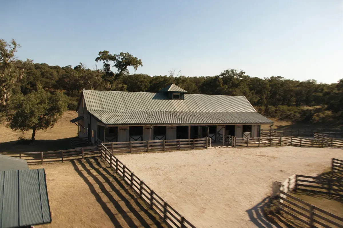 A center-aisle horse barn with a cupola and board-fenced paddocks in the Texas Hill Country